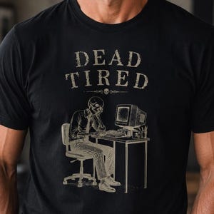May include: A black short-sleeved t-shirt featuring a beige graphic print of a skeleton sitting at a desk working on a computer. Above the illustration, the text reads DEAD TIRED in a distressed font, with a small skull icon centered below the text.