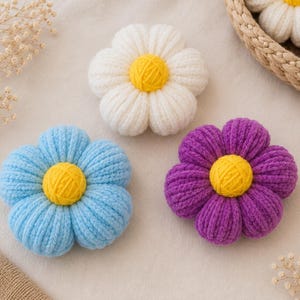 May include: Three plush flower-shaped items made of soft yarn. One flower is white, one is light blue, and one is purple, each featuring a bright yellow circular centre. The petals are rounded and textured, creating a soft, padded appearance.