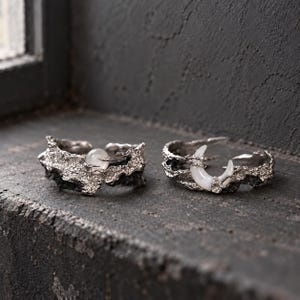 May include: Two silver-toned open rings with a rough, textured finish. One ring features a small, round, translucent white stone, while the other ring displays a crescent moon-shaped white stone set within the jagged metal band.