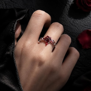 May include: A silver-toned ring featuring a central, irregular, deep red enamel accent that mimics a dripping liquid effect. The metal band has a textured, organic surface that flows into the red detail, creating a molten appearance on the finger.