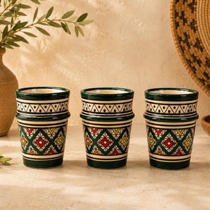 Puede incluir: Tres tazas cilíndricas de cerámica con una base de color verde oscuro y un borde horizontal prominente. El exterior está decorado con un patrón geométrico repetitivo de rombos en rojo, amarillo y verde, rematado por una banda de chevrón.