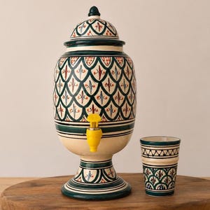 Puede incluir: Un dispensador de bebidas de cerámica con tapa y grifo de plástico amarillo, con una base color crema y patrones de escamas pintados en verde oscuro y rojo. A su lado hay un vaso de cerámica a juego con un motivo decorativo similar.