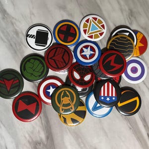 Puede incluir: Una colección de veinticuatro chapas redondas con diversos símbolos y emblemas gráficos coloridos. Los diseños incluyen iconos que representan un martillo, una estrella, un puño, una máscara de araña y patrones geométricos en rojo, azul, verde y dorado.