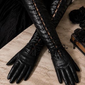 Puede incluir: Un par de guantes largos negros de estilo ópera con un diseño de cordones vertical que va desde la muñeca hasta el antebrazo. Los guantes tienen un acabado liso e incluyen finos cordones negros en la muñeca para un ajuste seguro.