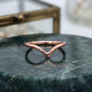 Pode incluir: Um anel em forma de V feito de metal de cor rosa com um acabamento martelado e texturizado. A banda apresenta uma silhueta de chevron distinta que termina em ponta no centro, refletindo a luz sobre a sua superfície metálica.