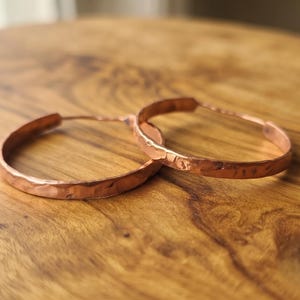 Könnte beinhalten: Ein Paar kreisförmige Creolen aus Kupfer mit einer ausgeprägten gehämmerten Textur auf der gesamten Oberfläche der Metallbänder. Die Ohrringe haben ein warmes, metallisch rotbraunes Finish und ein schlichtes, durchgehendes Ringdesign.