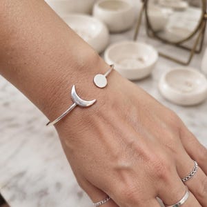 Könnte beinhalten: Ein silberfarbenes Armband in offener Form mit einem Mondsichel-Anhänger an einem Ende und einer kleinen runden Scheibe am anderen. Das Armband wird am Handgelenk getragen, ergänzt durch zwei schlichte silberfarbene Ringe, einer glatt und einer mit Kettengliedern.