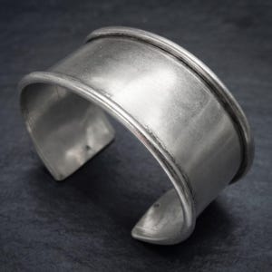 Puede incluir: Una pulsera brazalete ancha de metal plateado con una superficie lisa y pulida, con bordes elevados y redondeados en la parte superior e inferior. La pulsera presenta un diseño abierto en la parte posterior, mostrando una banda curva.