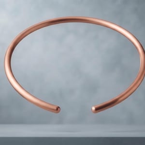 Può includere: Un bracciale rigido in metallo color rame, liscio e circolare, con finitura lucida e una piccola apertura nella parte inferiore. Il bracciale presenta un design semplice a fascia arrotondata continua, che mantiene uno spessore costante lungo tutta la circonferenza.