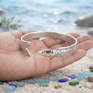 Puede incluir: Una pulsera tipo brazalete de metal plateado con forma de pez, que presenta un patrón de escamas texturizado a lo largo de la banda. La pulsera es abierta, con un extremo formando la cabeza del pez y el otro la aleta caudal, creando un diseño envolvente.