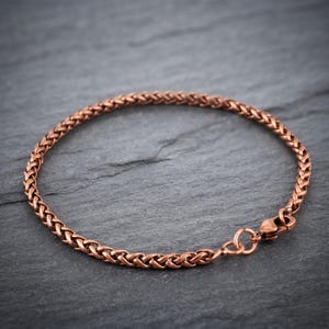 May include: A copper-toned wheat chain bracelet featuring a woven, braided metal link design. The bracelet is circular in shape and includes a small, functional lobster claw clasp mechanism at one end for securing the piece around the wrist.