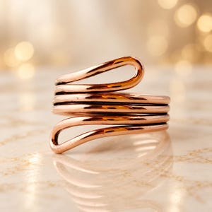 Puede incluir: Un anillo de cobre pulido con un diseño de alambre continuo que se envuelve varias veces. El metal tiene un tono naranja-marrón metálico cálido con una superficie lisa y reflectante. Los extremos del alambre son redondeados y forman bucles.