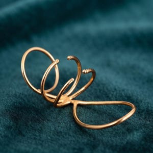 Può includere: Un anello in metallo color oro caratterizzato da un design a più fasce con tre anelli aperti e curvi che si estendono da una base singola. La struttura è composta da sottili segmenti metallici lucidi che creano un aspetto allungato.