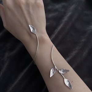 Peut inclure: Un bracelet de bras argenté composé d'une fine tige métallique incurvée qui s'enroule autour de l'avant-bras. Le design présente quatre petits éléments en forme de feuilles à la texture martelée fixés aux extrémités et le long de la tige.