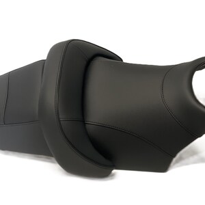 Peut inclure: Une selle de moto noire présentant un design en deux parties avec une section passager surélevée et une zone conducteur profilée. La selle est fabriquée dans un matériau lisse au fini mat et comporte des coutures visibles sur les bords.