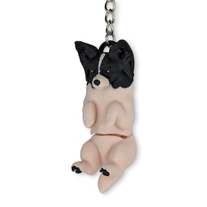 May include: A 3D printed keychain featuring a Papillon dog with black ears and a white face stripe, transitioning into a light beige body. The dog is depicted in a hanging position with front paws tucked and back paws extended, attached to a metal chain.
