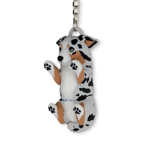May include: A keychain featuring a three-dimensional figurine of a dog with a white, black, and tan merle coat pattern. The dog is depicted in a seated position with its front paws raised, and it is attached to a silver-colored metal chain.