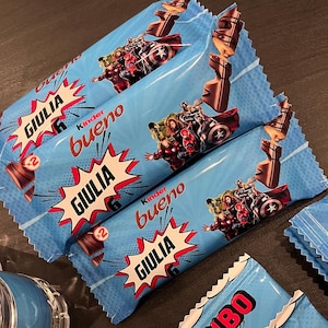 May include: Three light blue Kinder Bueno chocolate bar wrappers featuring Marvel superhero graphics, the Kinder Bueno logo, and the name GIULIA printed in a white comic-style speech bubble. Each wrapper displays images of chocolate bars.