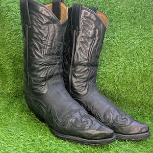 Op de afbeelding: Een paar zwarte leren cowboylaarzen met gedetailleerde ton-sur-ton stikselpatronen op de schacht en de wreef. De laarzen hebben een puntige neus, een hak en treklussen aan de bovenkant van de schacht, met een beige voering bij de opening.