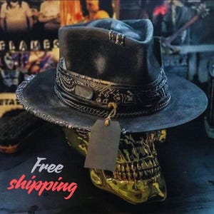 May include: A black fedora hat featuring a wide brim and a crown wrapped in a black and white patterned bandana. A thin metal chain encircles the base of the crown, and a blank rectangular tag hangs from the front of the hat by a brown string.