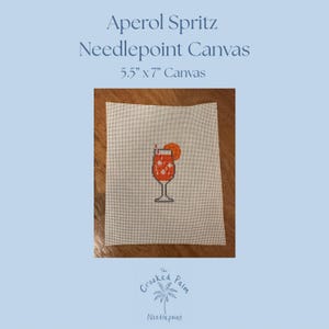 May include: A white needlepoint canvas measuring 5.5 inches by 7 inches, featuring a centered graphic of an Aperol Spritz cocktail in a glass with an orange slice garnish. The text Aperol Spritz Needlepoint Canvas and The Crooked Palm Needlepoint appear.