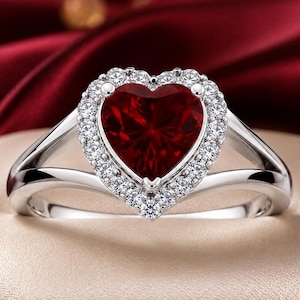 May include: A silver ring featuring a large, heart-shaped deep red gemstone at its centre. The heart stone is surrounded by a halo of small, clear, round-cut white stones. The band is made of polished silver and splits into two sections.