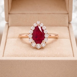 Peut inclure: Une bague avec une pierre rouge en forme de poire montée sur un anneau en or rose. La pierre centrale est entourée d'un halo de petites pierres blanches rondes et transparentes. La bague est présentée dans un écrin beige clair.