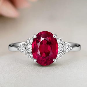 Puede incluir: Un anillo de plata con una gran piedra preciosa de color rojo intenso y corte ovalado en el centro, sujeta por cuatro garras. El aro está adornado a cada lado con un grupo de tres pequeñas piedras redondas, claras y brillantes.
