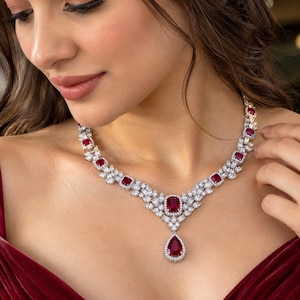 May include: A necklace featuring a central pear-shaped red gemstone drop, surrounded by smaller cushion-cut red stones and numerous clear white stones set in a metallic frame, creating a symmetrical arrangement that rests along the collarbone.