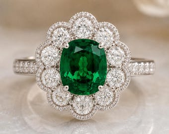 Puede incluir: Un anillo de tono plateado con una gran piedra central de color verde intenso y corte ovalado, rodeada por un halo de diez piedras redondas transparentes de talla brillante. El aro y el engaste festoneado tienen pequeñas piedras.