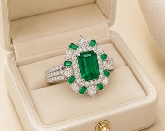 Puede incluir: Un anillo de tono plateado con una gran piedra central verde de corte esmeralda rodeada por un halo de piedras blancas más pequeñas y piedras verdes de corte baguette. La banda se divide en tres filas incrustadas con pequeñas piedras blancas.