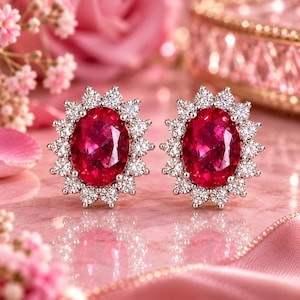 May include: A pair of oval-shaped stud earrings featuring a vibrant red centre stone surrounded by a halo of small, clear, round-cut white stones. The setting is metallic, and the design creates a floral-like appearance with faceted surfaces.