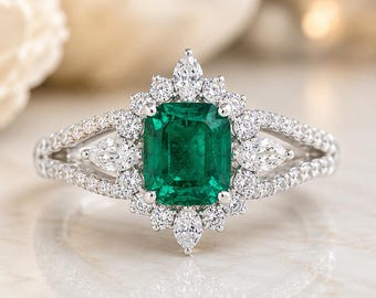 Puede incluir: Un anillo de tono plateado con una piedra preciosa verde central de corte esmeralda, rodeada por un halo de pequeñas piedras claras redondas y de corte marquesa. La banda dividida está incrustada con diminutas piedras claras.