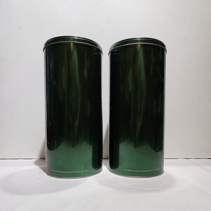 以下が含まれることがあります： 丸みを帯びた蓋が付いた、2 つの背の高い円筒形の濃い緑色の金属容器。容器は光沢仕上げで、並んで配置されています。各容器の底にはテキストが印刷されています。