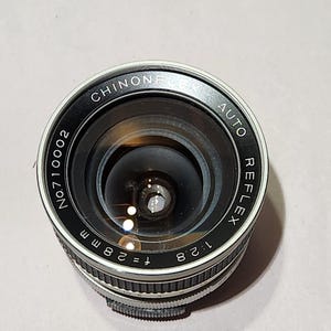 May include: A black and silver Chinon Auto Reflex camera lens. The lens has the text "CHINON" and "AUTO REFLEX" printed on the outer ring. The lens has a focal length of f=28mm and an aperture of 1:2.8.