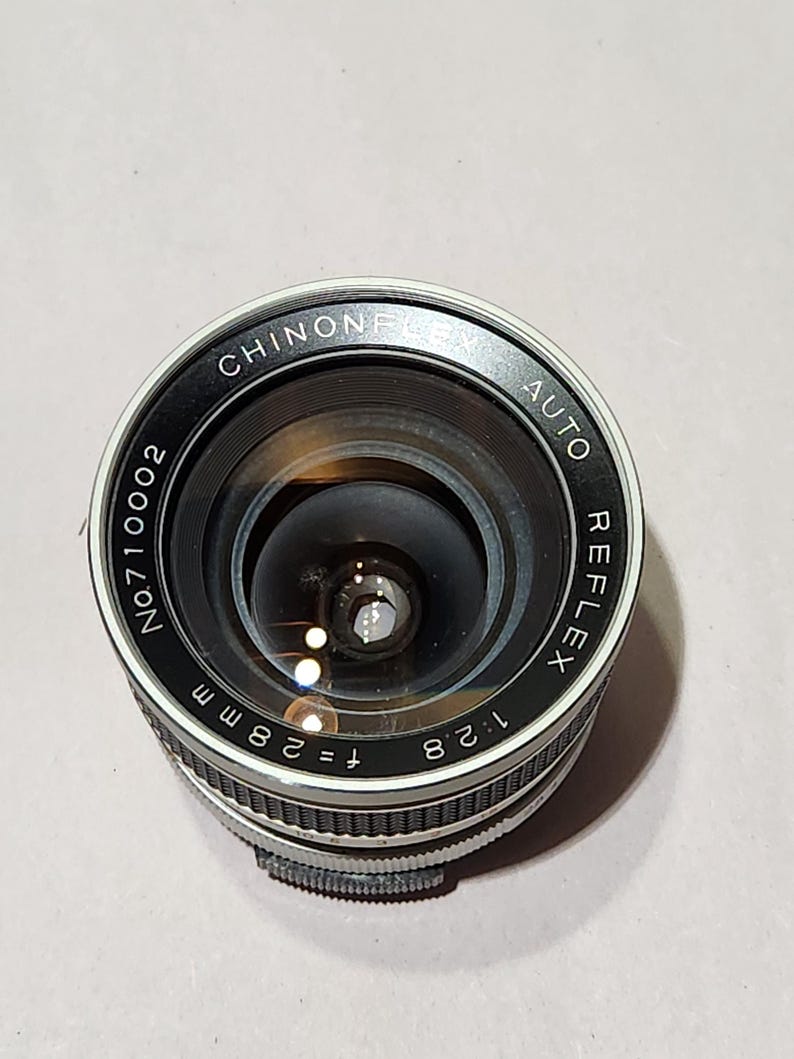 May include: A black and silver Chinon Auto Reflex camera lens. The lens has the text "CHINON" and "AUTO REFLEX" printed on the outer ring. The lens has a focal length of f=28mm and an aperture of 1:2.8.