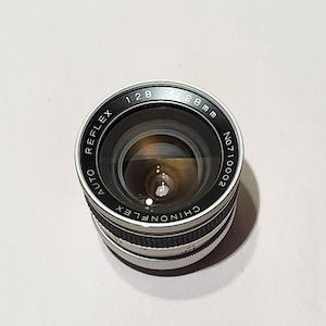 May include: A Chinonflex Reflex Auto 28mm camera lens with a silver and black body. The lens has a 1:2.8 aperture and is marked with the serial number No.710002. The lens is designed for use with cameras.