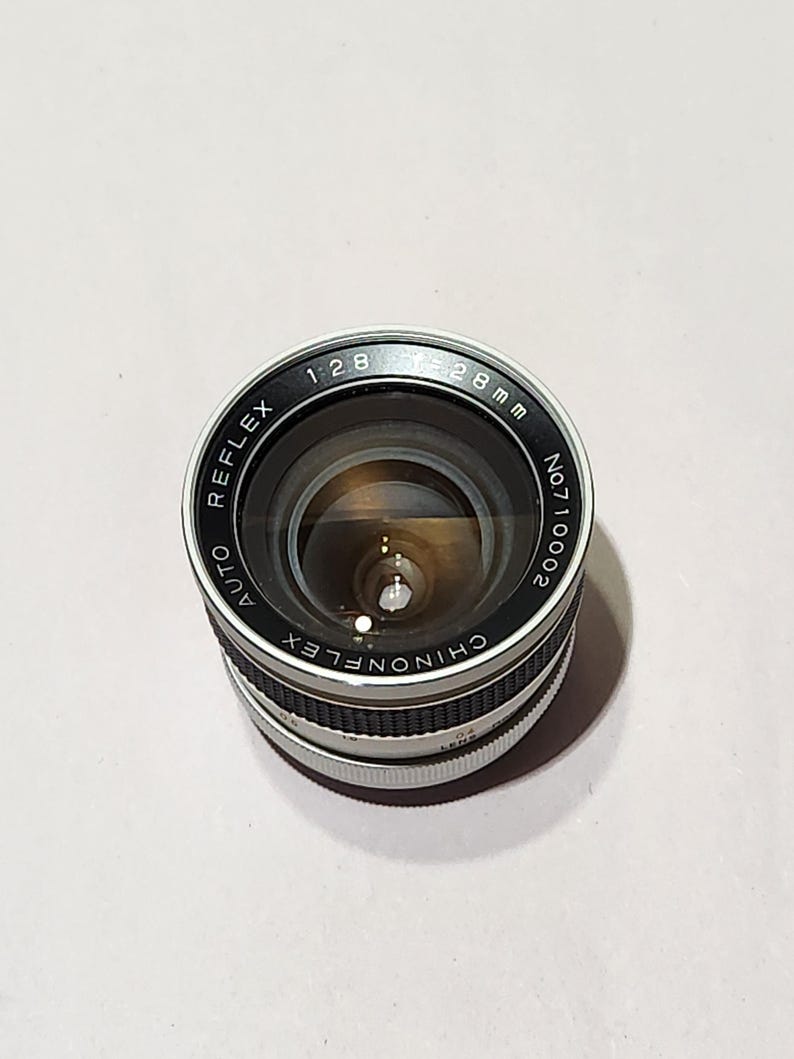 May include: A Chinonflex Reflex Auto 28mm camera lens with a silver and black body. The lens has a 1:2.8 aperture and is marked with the serial number No.710002. The lens is designed for use with cameras.