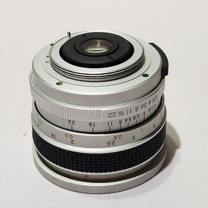 May include: A silver camera lens with black rubber grip and focus ring. The lens has a series of numbers and markings, including f-stops and distance measurements. The lens is designed for a camera.