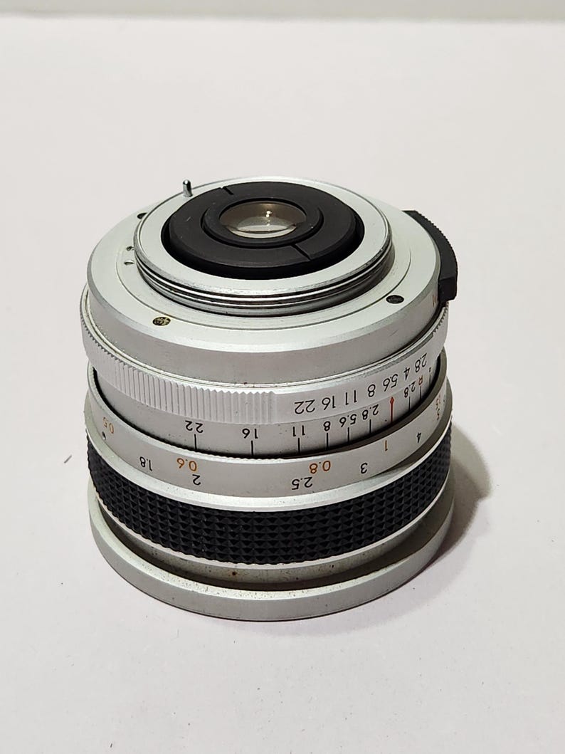 May include: A silver camera lens with black rubber grip and focus ring. The lens has a series of numbers and markings, including f-stops and distance measurements. The lens is designed for a camera.