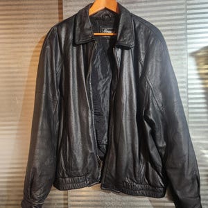 Peut inclure: Une veste en cuir noir dotée d'un col classique, d'une fermeture éclair frontale intégrale et de manches longues avec des poignets côtelés. La veste comprend un ourlet côtelé et une doublure intérieure noire, avec une étiquette de marque visible.