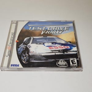 Op de afbeelding: Een Sega Dreamcast-gamecase voor Test Drive V-Rally. De case is van doorzichtig plastic met een wit label waarop een witte auto met rode en blauwe accenten te zien is. De auto heeft het woord "ESSO" op de voorkant. Het label heeft ook de woorden "Test Drive V-Rally" en "Sega Dreamcast" erop.