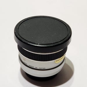 May include: A vintage camera lens with a black cap. The lens has a white and silver body with the text "MADE IN JAPAN". The lens is cylindrical and has a ridged grip. A sticker on the lens reads "PASSED".
