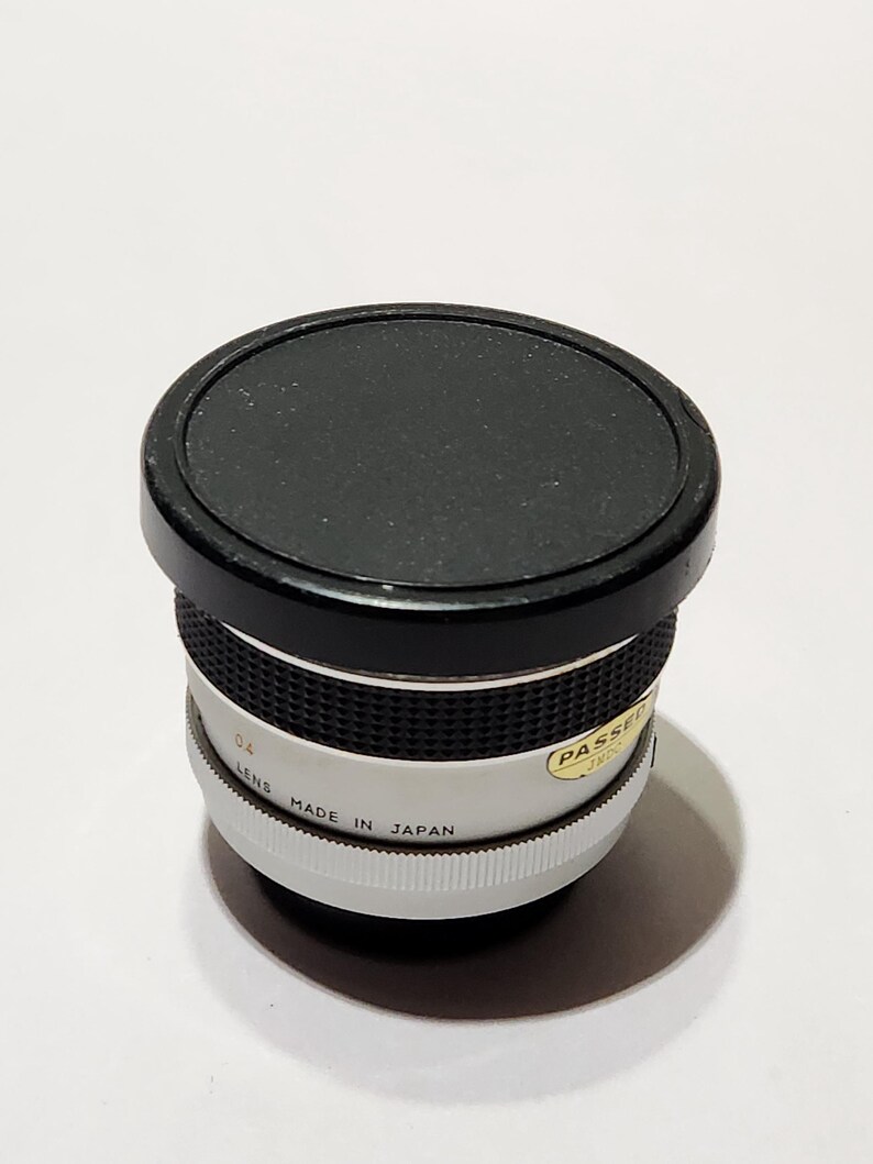 May include: A vintage camera lens with a black cap. The lens has a white and silver body with the text "MADE IN JAPAN". The lens is cylindrical and has a ridged grip. A sticker on the lens reads "PASSED".