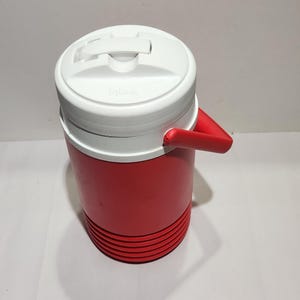 May include: A cylindrical red plastic beverage cooler with a white screw-on lid featuring a molded handle and the Igloo brand name embossed on the top. The base of the cooler has a series of horizontal ridges, and a red side handle is attached.