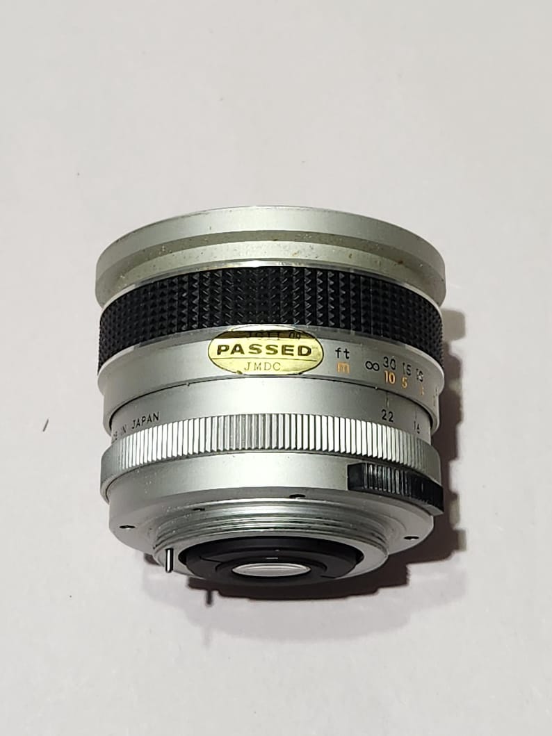May include: Silver and black camera lens with a ridged focus ring and aperture settings. The lens has a "PASSED" sticker and markings in feet and inches. The lens is marked "MADE IN JAPAN".