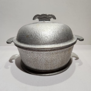 May include: A silver-colored metal casserole dish with a textured, hammered finish. The round pot features two side handles and a matching domed lid with a decorative, scalloped handle on top. The exterior surface has a consistent, rough, pebbled appearance.