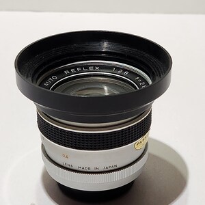 May include: A vintage camera lens with a black lens hood and a silver and black body. The lens is marked "AUTO REFLEX 1:2.8 f=28" and "LENS MADE IN JAPAN". The lens has a 0.4 measurement.