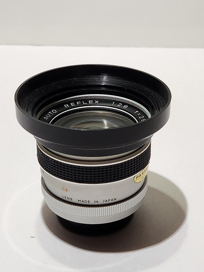 May include: A vintage camera lens with a black lens hood and a silver and black body. The lens is marked "AUTO REFLEX 1:2.8 f=28" and "LENS MADE IN JAPAN". The lens has a 0.4 measurement.