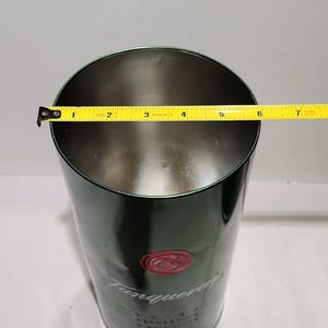 以下が含まれることがあります： タンカレーのロゴと赤いエンブレムが付いた、濃い緑色の金属製アイスバケツ。バケツは円筒形で、直径は約15cmです。黄色の巻尺が上部に置かれています。